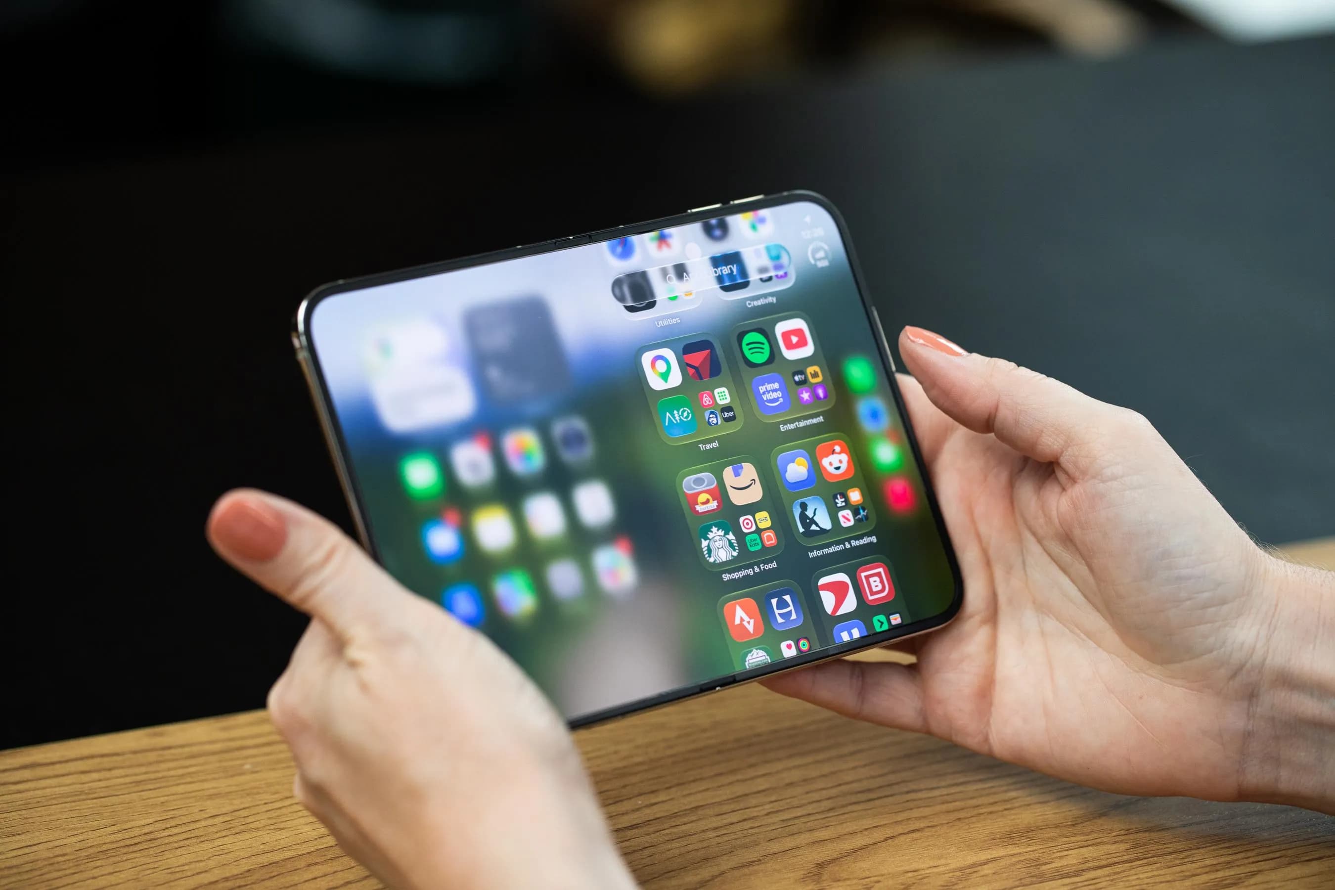 Hands holding the iPhone Duo open to the inner screen in landscape orientation. The left side of the screen is blurred out, while the right side shows the app library.