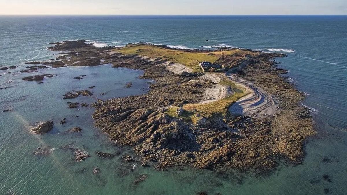 An aerial photograph shows a small rocky island surrounded by clear blue-green sea, with a patchwork of grassland, rugged shoreline and a handful of buildings near the centre connected by narrow tracks.