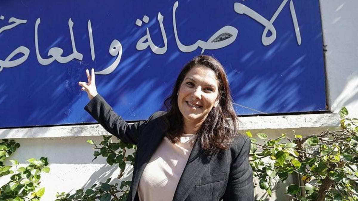 El Mansouri, wearing a black blazer over a white shirt, stands in front of her party's slogan, written in Arabic in white lettering against a blue background.