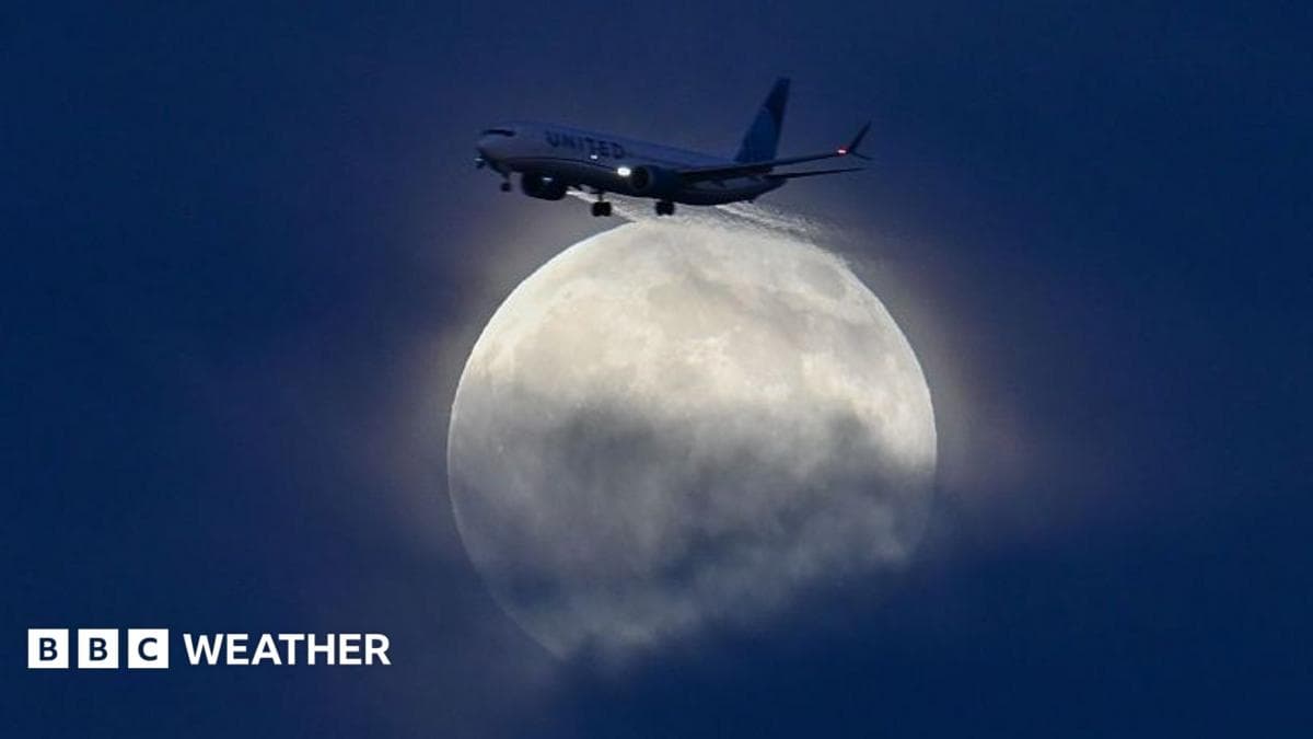 A large, glowing white moon is rising through blue cloud as a plane flies. The plane is at the top of the moon and the sky is a dark blue behind it.
