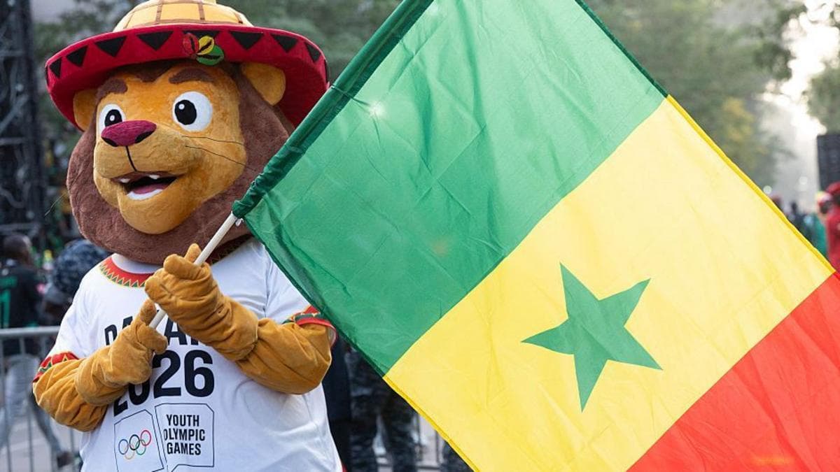 A friendly-looking lion mascot holding up a large national flag of Senegal on a pole. The mascot, called Ayo, wears a colourful red-and-yellow traditional-style hat and a white T-shirt featuring the logo for the Dakar 2026 Youth Olympic…