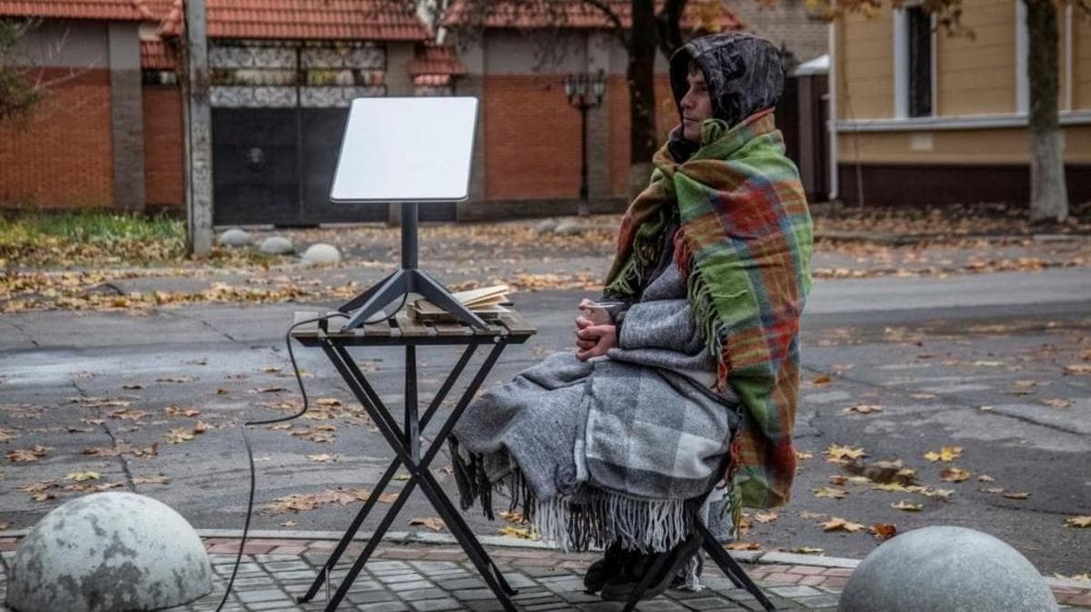 A man, with at least three blankets around him and a drink in his hand, sits on a chair on the side of a street. Next to him is a small picnic table with a starlink terminal on it.
